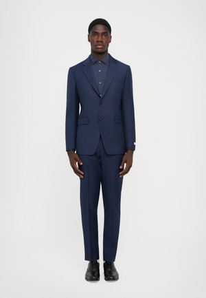 Marineblauw op maat gemaakt pak met een blazer met één knoop en bijpassende broek. Gelaagde stof met subtiele glans, gedragen over een donker button-up shirt.