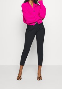 Cárdigan rosa con canalé y botones, combinado con pantalones negros a medida. La modelo lleva sandalias negras de dedo, posando frente a un fondo liso.