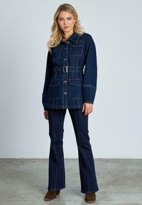 Giacca di jeans scuri con vita a cinturino, chiusura a bottoni, quattro tasche e cuciture a contrasto. Abbinata a jeans svasati scuri e stivali alla caviglia.