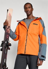 Άνδρας που φοράει πορτοκαλί και γκρι μπουφάν σκι και μαύρο παντελόνι, κρατώντας ένα ζευγάρι σκι κατακόρυφα σε ουδέτερο φόντο.