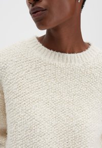 Gebreide crème trui met een gestructureerd, bouclé patroon, voorzien van een geribde ronde hals. De stof lijkt zacht en chunky te zijn.