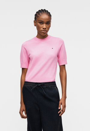 Woman with braided hair wearing a fitted pink short-sleeve shirt and black drawstring pants, standing against a plain light background.