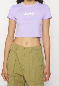 Levandulový crop top s krátkými rukávy a vlnitým lemem. Obsahuje bílý nápis "obey". V páru s olivově zelenými cargo kalhotami.