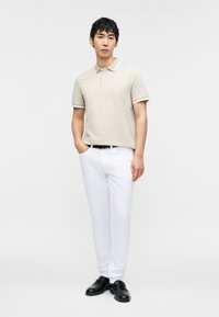 Ανοιχτό μπεζ πόλο πουκάμισο με διακριτικό μοτίβο, με γιακά και μανσέτες με λευκή περιθώρια. Συνδυασμένο με λευκό παντελόνι και μαύρα παπούτσια.