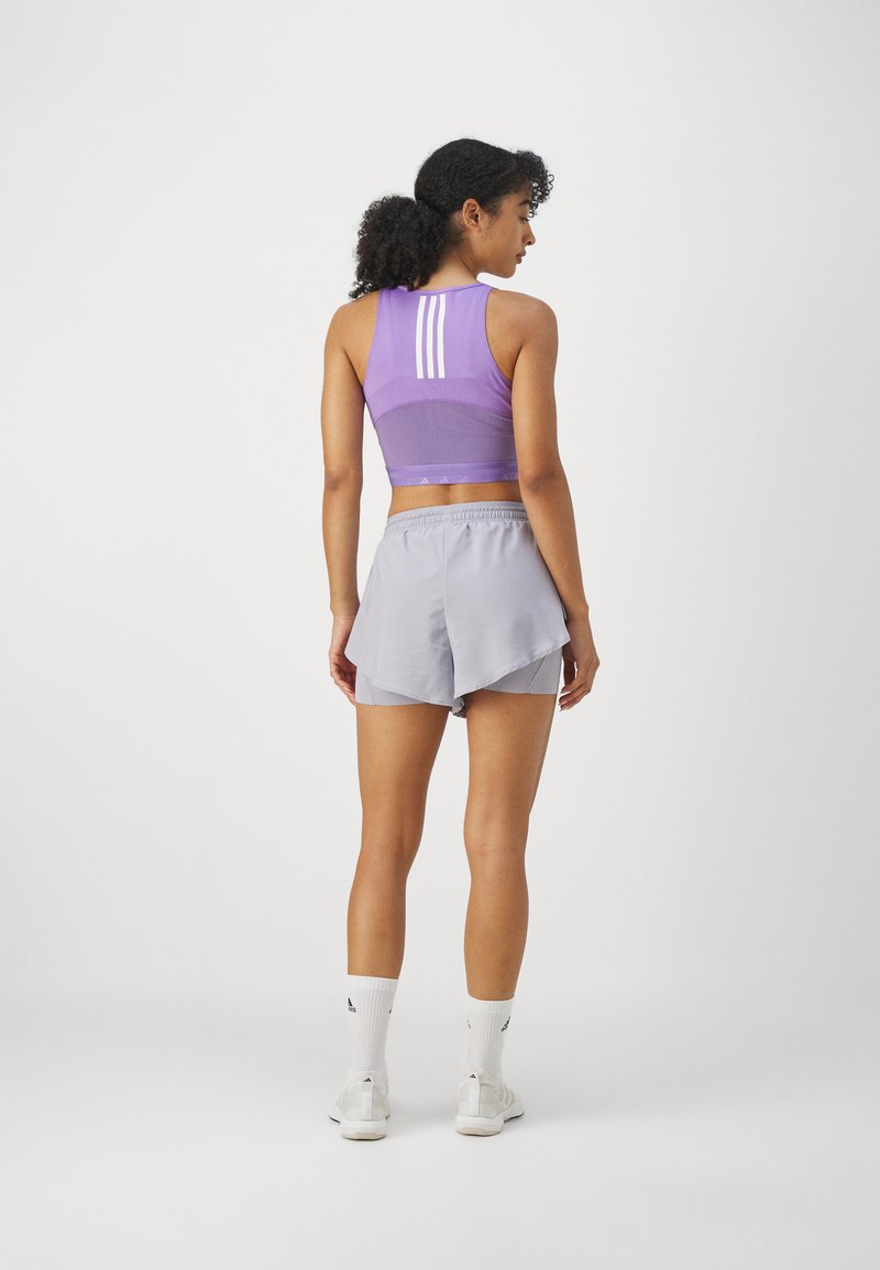 Sujetador deportivo morado con tres rayas blancas en la parte posterior, combinado con pantalones cortos gris claro. El material parece elástico y transpirable.