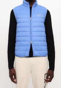 Světle modrá pufovaná vesta s vertikálním prošíváním a stojáčkovým límcem. Obsahuje přední zip a logo EA7 na hrudi.