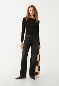 Schwarzes langärmliges Oberteil, hochgeschnittene, dunkelblaue, weite Jeans, spitze schwarze Ballerinas mit Nieten, hält einen braun-weißen karierten Schal.