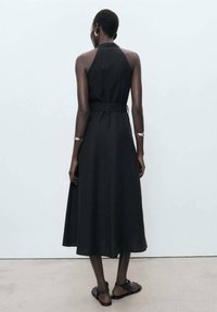 Black, sleeveless midi dress with a belted waist and a halter neckline. Smooth fabric with a flared hem, worn with flat sandals.