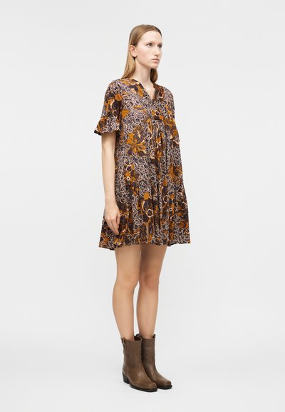 Γυναίκα που στέκεται, φοράει κοντό λουλουδάτο φόρεμα με φουσκωτά μανίκια και καφέ μποτάκια αστραγάλου, σε απλό λευκό φόντο.