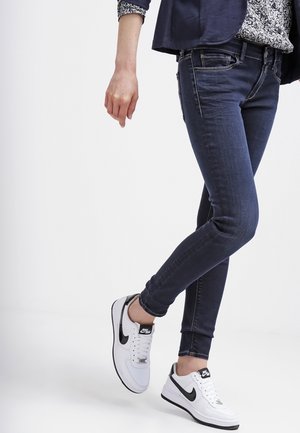 Osoba ubrana w ciemne wąskie dżinsy i białe sneakersy Nike Air, z jedną nogą założoną na drugą na prostym tle.