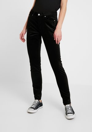 Kvinde iført sorte slim-fit jeans og sorte lavtop sneakers med hvide snørebånd, stående mod en ensfarvet lys baggrund.