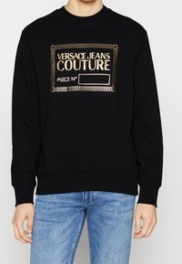 Svart sweatshirt i bomull med ribbat hals- och ärmslut, med en broderad logopatch i guld på framsidan.