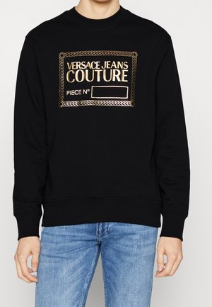 Sort sweatshirt lavet af bomuld med ribstrikket halsudskæring og manchetter, med et guld broderet logo-patch på forsiden.