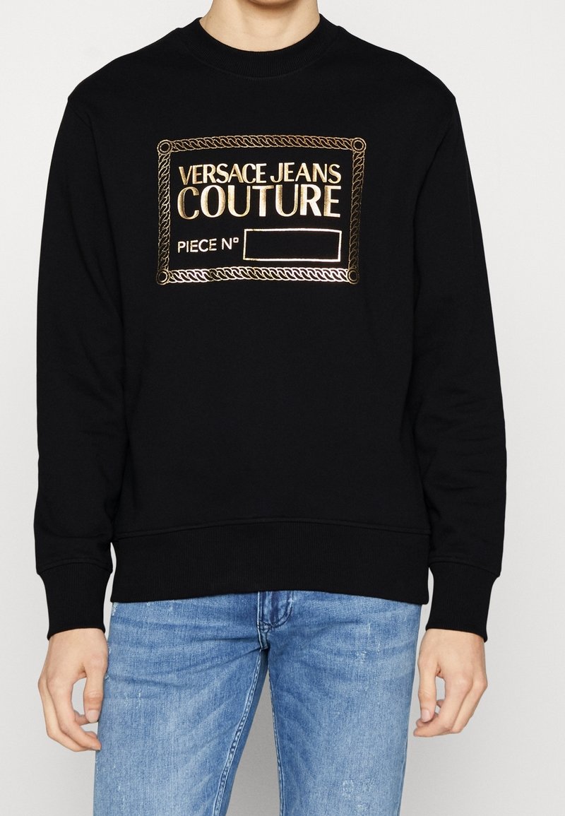 Svart sweatshirt i bomull med ribbat hals- och ärmslut, med en broderad logopatch i guld på framsidan.