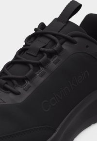 Gros plan d'une basket noire Calvin Klein montrant des lacets épais, des passants de lacets et le nom de la marque en relief sur le côté.