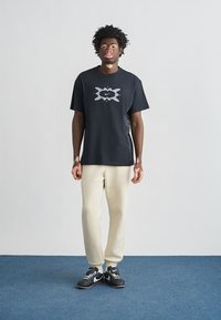 Μαύρο κοντομάνικο μπλουζάκι με κεντρικό γκρι λογότυπο της Nike, σε συνδυασμό με παντελόνι σε κρεμ απόχρωση και μαύρα αθλητικά παπούτσια σε μπλε πάτωμα.