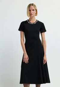 Vestido negro hecho de tela suave, con mangas cortas y dobladillo acampanado. Escote adornado con detalles de cuentas reflectantes.
