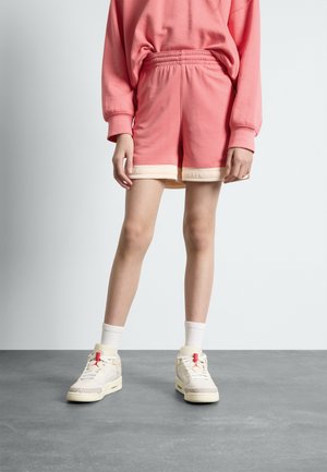 Lyserøde teksturerede shorts med en kontrasterende cremefarvet talje, parret med cremefarvede sneakers med grå accenter og røde detaljer.