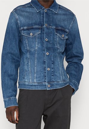 Homme portant une veste en jean bleu moyen avec des boutons à l'avant et des poches poitrine, associée à un pantalon foncé, debout avec une main dans la poche.