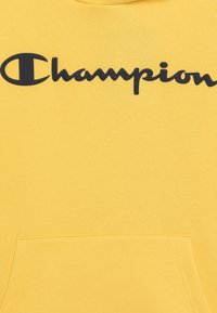 Felpa gialla con tasca frontale, caratterizzata da un grande logo nero "Champion" sul petto. Tessuto morbido con una superficie liscia.