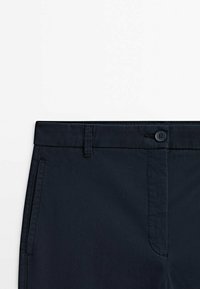 Détail de la taille d'un pantalon bleu marine foncé montrant la fermeture par bouton, les passants de ceinture et les poches latérales sur un fond blanc.
