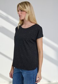 Heledate juustega naine, kes kannab musta lühikeste varrukatega särki ja siniseid teksaseid, seisab siseruumis valge seinaga, millel on diagonaalsed varjud.