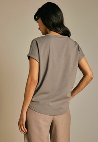 T-shirt en coton gris avec des manches courtes roulées et un ourlet arrondi, porté avec un pantalon beige clair, présenté sur un fond neutre.