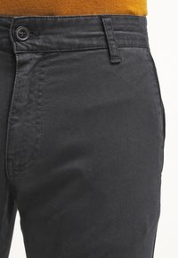 Pantalon gris foncé en tissu lisse, doté d'une fermeture à bouton et d'un devant plat. Couture visible le long des coutures.