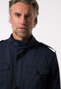 Námořnická modrá bunda s vysokým límcem, zapínáním na knoflíky a dvěma náprsními kapsami. Vyrobena z hladké tkaniny s ramenními epauletami.