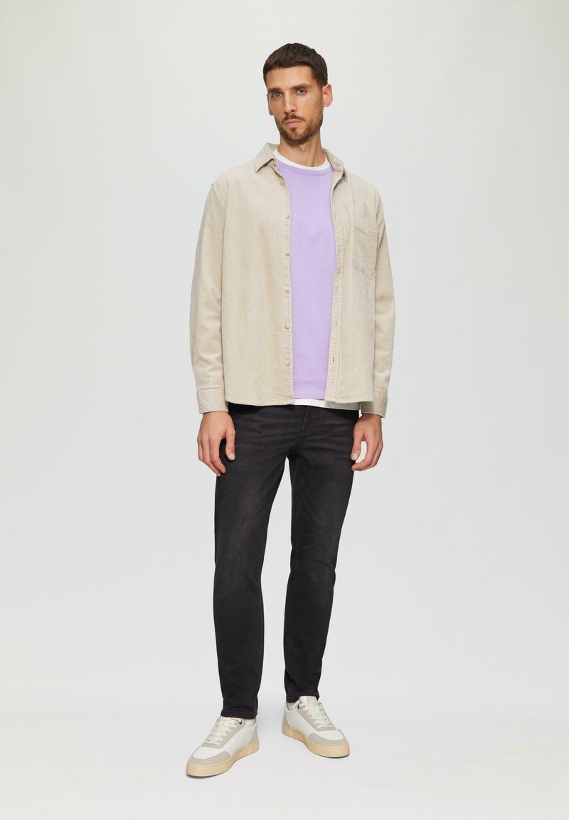 Hellbeige Cord-Shirts über einem lila T-Shirt, kombiniert mit schmalen schwarzen Jeans und cremefarbenen Sneakers mit beigen Akzenten. Schlichte Hintergrund.