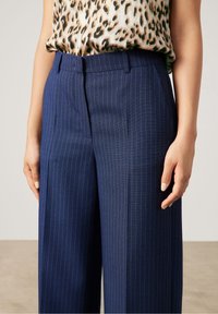 Marineblå skräddarsydda byxor med vertikala vita ränder, med en slät front, sidofickor och en formsydd midja. Slät tygtextur.