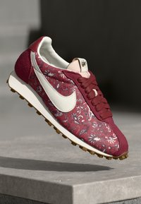 Crvene tenisice s cvjetnim uzorkom, bijeli Swoosh logo, teksturirana gornja površina, padding oko zapešća i gumeni potplat s bež profilom.