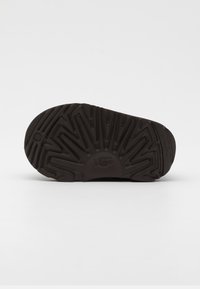 Semelle de chaussure en caoutchouc noir avec un motif de grip texturé, présentant des rainures triangulaires et un détail de marque circulaire près du talon.