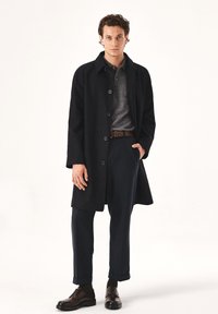 Abrigo de lana negro con corte recto y botones grandes, llevado sobre un suéter gris y pantalones oscuros. Zapatos marrones con cordones completan el conjunto.