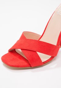 Sandale rouge en daim à bout ouvert avec des brides croisées et une semelle intérieure beige sur fond blanc.
