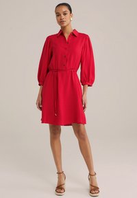 Robe rouge jusqu'aux genoux avec un devant à boutons, des manches ballons courtes et une taille à cordon. Fabriquée en tissu texturé.