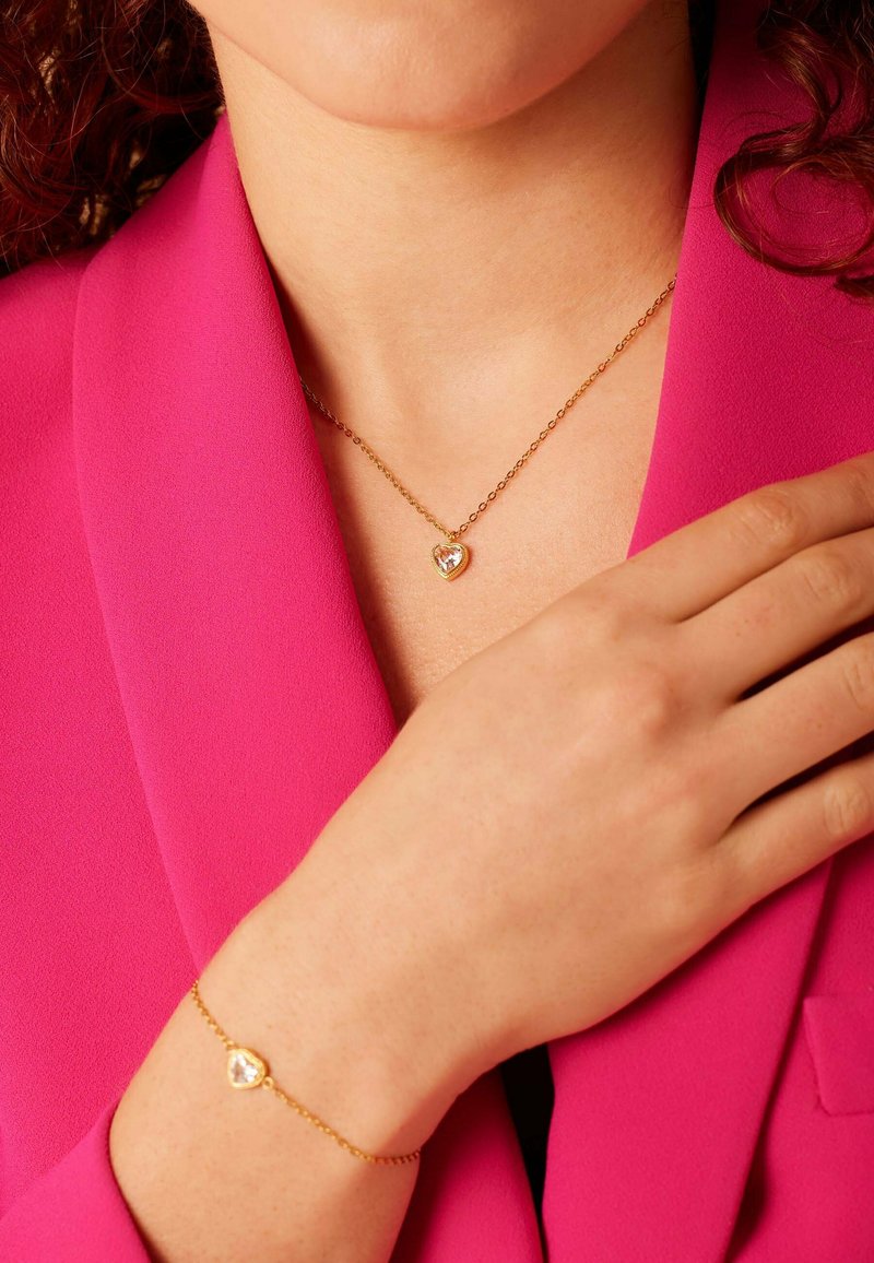 Gouden hartvormige ketting met heldere steen, gelaagd over een roze blazer. Gouden armband met bijpassend hartdetail om de pols.