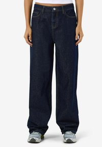 Osoba u tamnoplavim širokim trapericama i sivim tenisicama, stoji pred jednostavnom bijelom pozadinom.