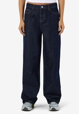 Oseba v temno modri širokih hlačah in sivih supergah, stoji ob proti enobarvni beli ozadju.