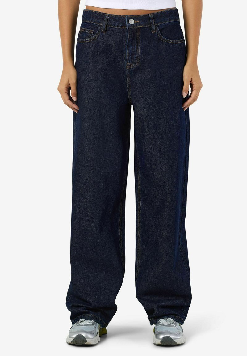 Osoba u tamnoplavim širokim trapericama i sivim tenisicama, stoji pred jednostavnom bijelom pozadinom.