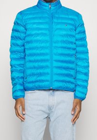 Giacca puffy azzurro chiaro con cuciture orizzontali, colletto rialzato, zip completa e tasche laterali. Il materiale appare lucido e leggero.