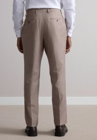 Pantalons beiges avec une texture lisse, dotés de deux poches arrière avec boutons, coupe ajustée et pli sur le devant ; associés à des chaussures noires.