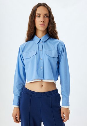 Camisa de manga larga recortada de algodón azul claro con botones al frente, dos bolsillos en el pecho y dobladillo blanco contrastante, combinada con pantalones elásticos de cintura navy.