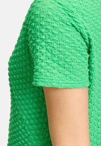 Grünes strukturiertes Oberteil mit Waffelstrickmuster, kurzen Ärmeln und einem weichen, atmungsaktiven Material. Die Nahtdetails sind an der Schulter sichtbar.