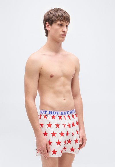 Άσπρα ανδρικά μποξεράκια με κόκκινα αστέρια, ελαστική μέση με μπλε κείμενο "HOT HOT HOT". Το μοντέλο στέκεται πλάγια, γυμνό το πάνω μέρος του σώματος, ουδέτερο φόντο.