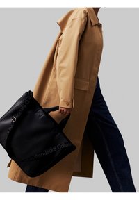 Vaaleanruskea trench-coat tummien farkkujen päällä, kannettuna mustan nahkaisen olkalaukun kanssa, jossa on kohokuvioitu logo ja teksturoitu pinta, tyylikkäällä suunnittelulla.