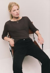 Rudas ilgomis rankovėmis, gofruotas megztinis derinamas su juodomis korduro švarkuota kelnėmis. Modelis sėdi kėdėje, dėvi auksinius papuošalus ir turi subtilią išraišką.