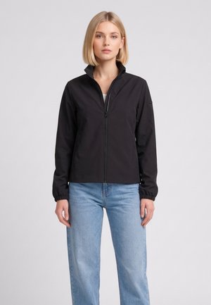 Donna bionda che indossa una giacca nera con cerniera e jeans azzurri, in piedi davanti a uno sfondo grigio uniforme.