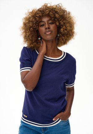 Kobieta o kręconych włosach, ubranа w granatowy sweter z krótkim rękawem z białymi paskami, niebieskie dżinsy i duże wiszące kolczyki, pozująca z ręką na szyi.