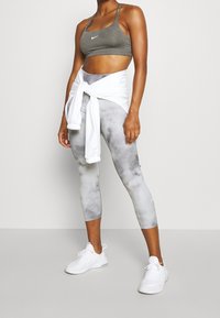 Vrouw die een grijze sportbeha, tie-dye cropped legging, witte sneakers en een wit jack om haar middel geknoopt draagt, staand tegen een effen achtergrond.